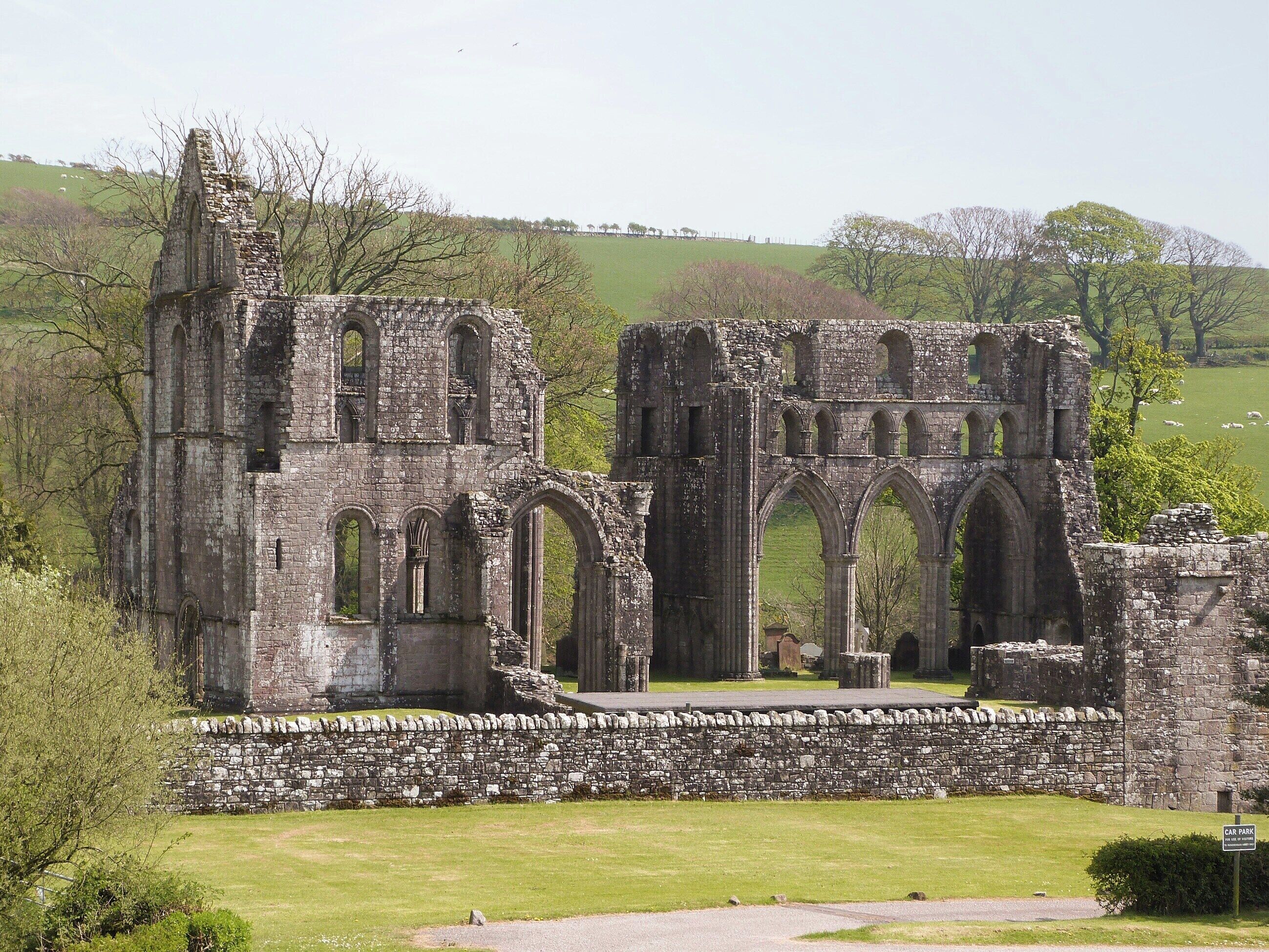 An ancient stone structure with multiple arches and a tower, surrounded by a stone wall and grassy area.