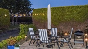 An outdoor patio with four striped chairs, a white umbrella, a barbecue grill, and string lights. - Hameish Holiday Cottage (Kirkcudbright)