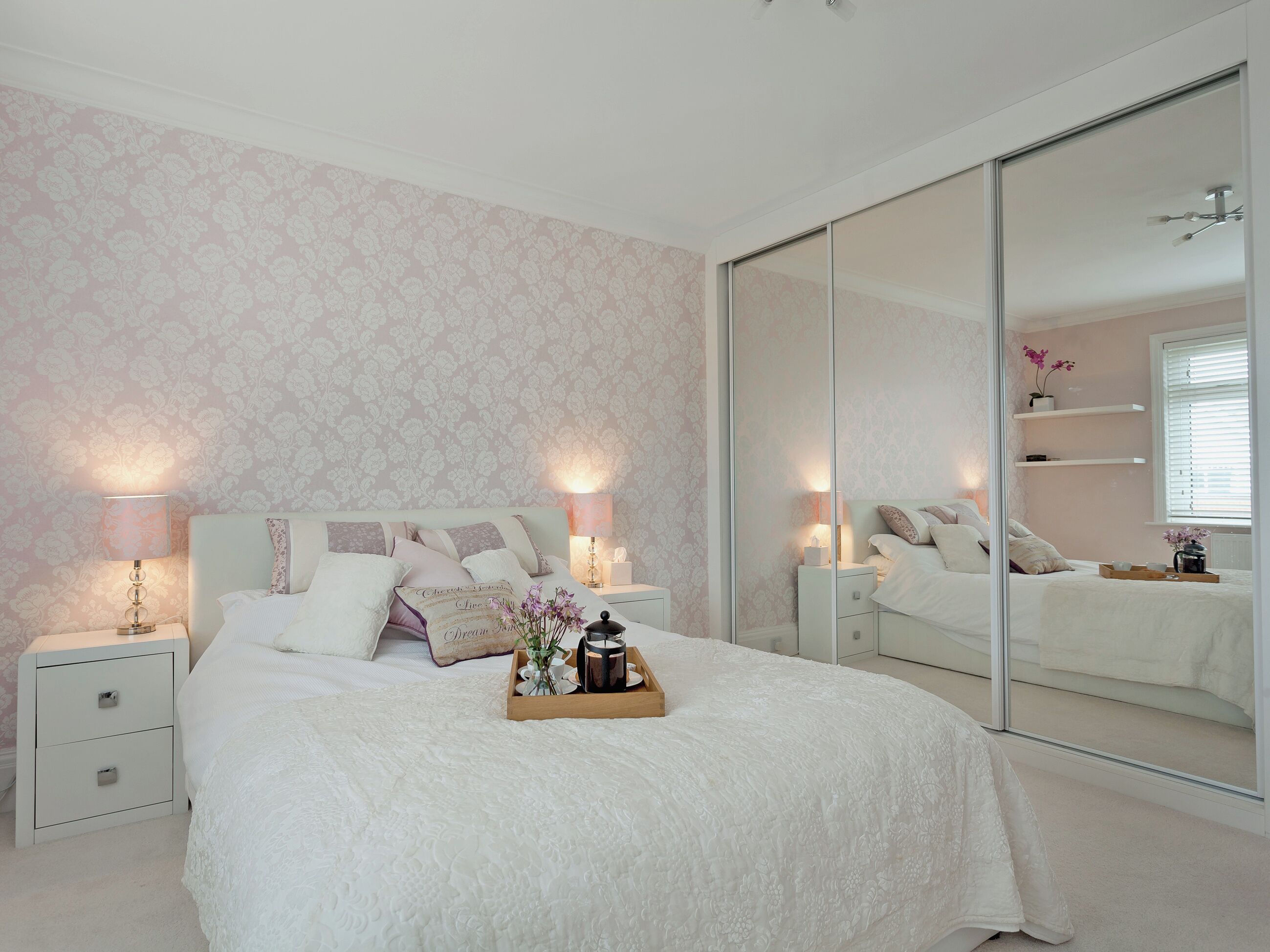 Un dormitorio con un espejo grande, una cama con ropa de cama blanca, mesitas de noche con lámparas y una bandeja con objetos sobre la cama.
