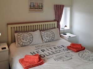 A neatly made bed with striped bedding and pillows, a bedside table with a lamp, a window with checkered curtains, and a wall-mounted artwork.
