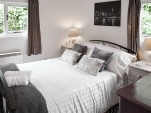 A neatly made bed with white bedding and multiple pillows, a bedside table with a lamp, and a window with curtains.