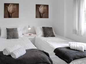 Une chambre d’hôtel avec deux lits, du linge de lit blanc, des oreillers gris, une table de chevet avec une lampe et deux tableaux représentant des fleurs, accrochés au mur.