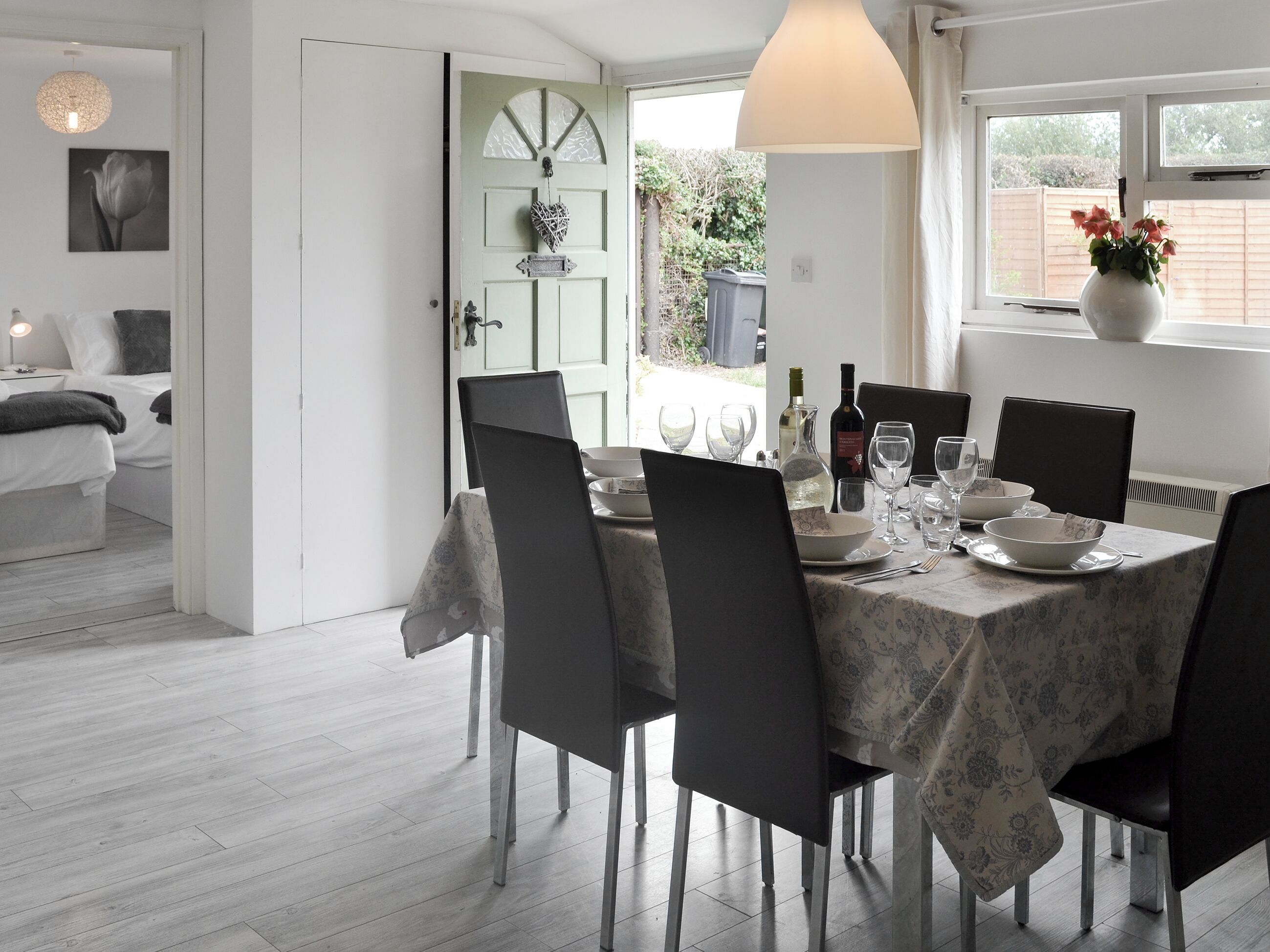 A dining area with a table set for six, chairs, a vase with flowers, and a window with a view of greenery.