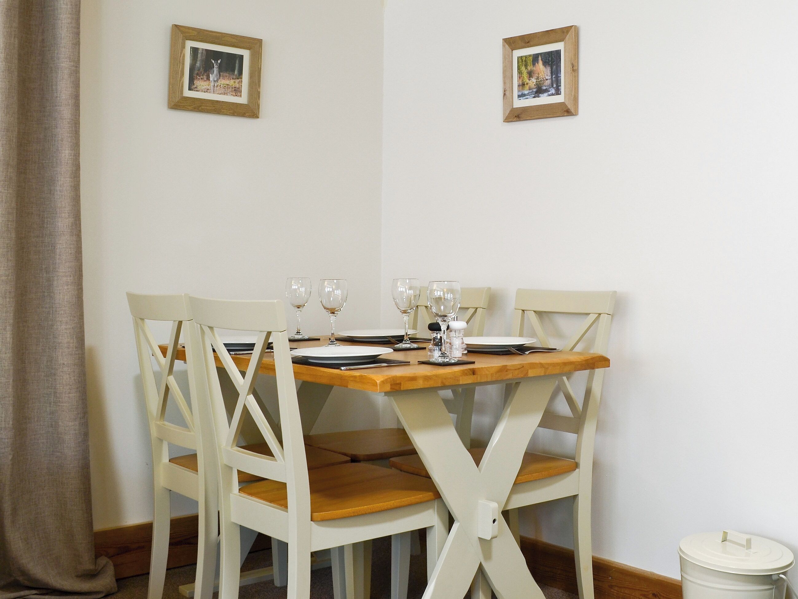 Une table à manger dressée pour quatre personnes, avec des chaises blanches, deux tableaux encadrés au mur et une poubelle placée sur le côté.