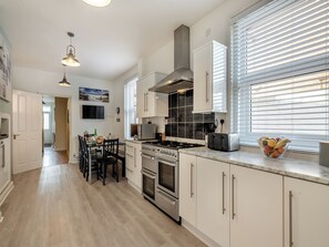 A modern kitchen with white cabinets, a gas stove, an oven, a microwave, and a toaster. There is a dining table with chairs and a window with blinds.