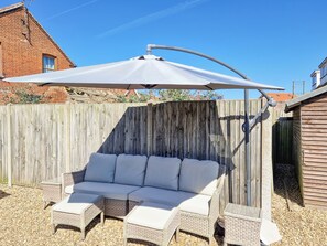 A covered outdoor seating area with a sofa and chairs, a wooden fence, and a building in the background.