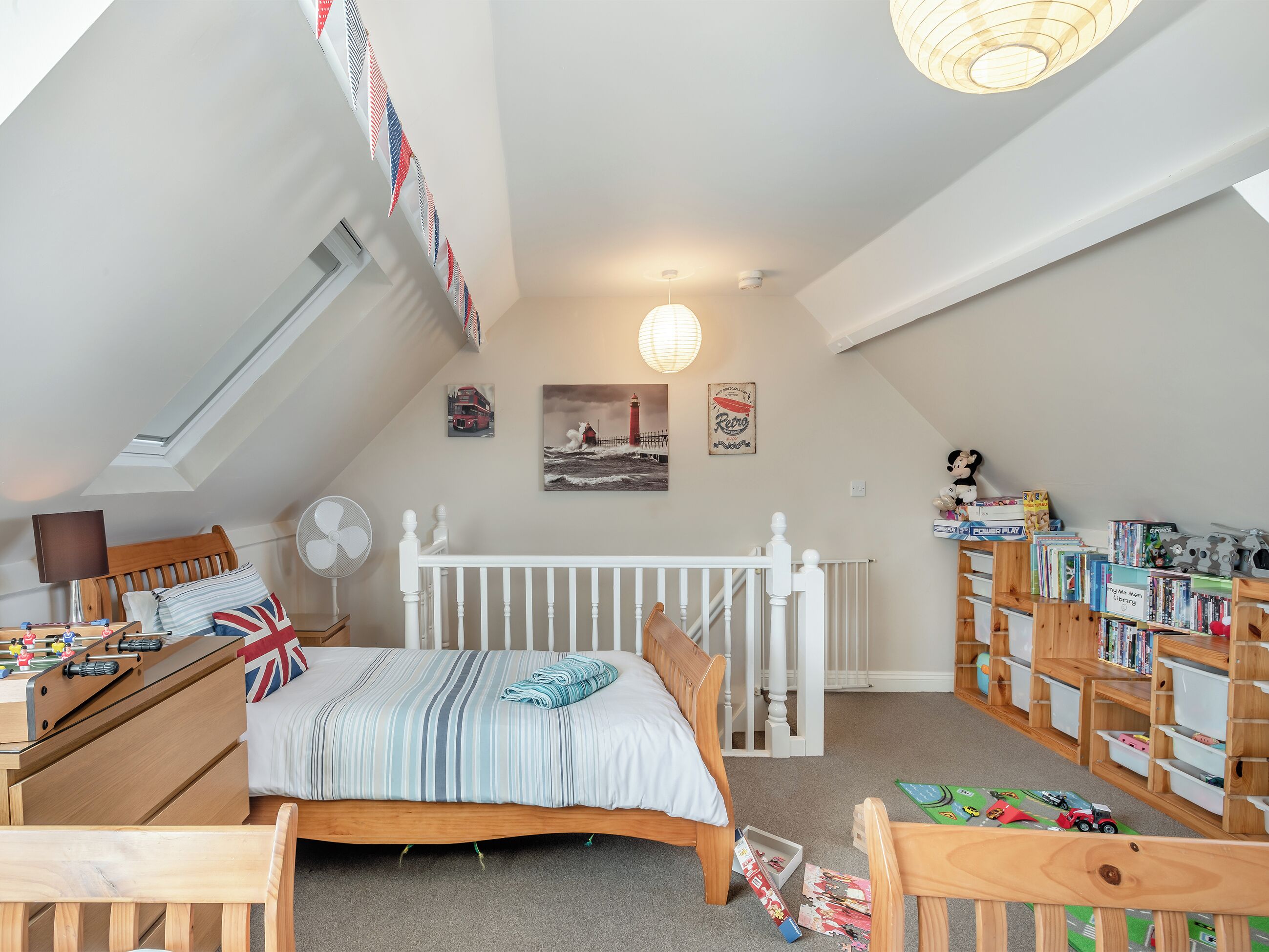 Un acogedor dormitorio en el ático, con techo inclinado, una cama con manta a rayas, un ventilador, una mesita con lámpara y una estantería llena de libros y juguetes.