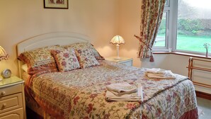 A bedroom with a floral bedspread, a bedside lamp, a framed picture on the wall, and a window with curtains.
