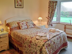 A bedroom with a floral bedspread, a bedside lamp, a framed picture on the wall, and a window with curtains. - South Cleeve Bungalow (Churchinford, near Taunton)