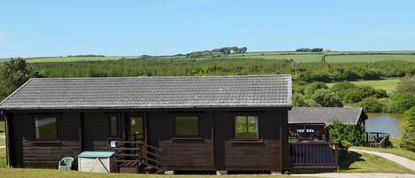 A wooden cabin with a porch and a small deck, surrounded by a grassy area and a body of water.