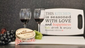 Un plan de travail de cuisine avec une grande meule de fromage, deux verres à vin et une pancarte sur laquelle on peut lire : « CETTE CUISINE est préparée avec amour et bonheur. Mangez, buvez et savourez. Tout est fait maison, avec amour. »