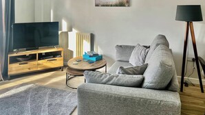 Un salon avec un canapé gris, un téléviseur posé sur un meuble en bois, une petite table basse ronde et un lampadaire.