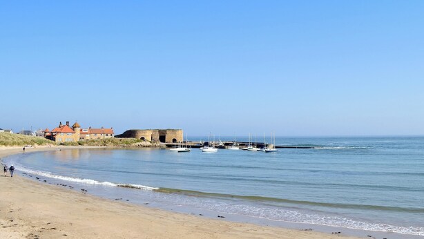 Un paysage côtier avec une plage de sable, un bâtiment historique et plusieurs bateaux ancrés près du rivage.