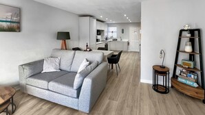 Un salon moderne avec un canapé gris, une table basse en bois et un fauteuil noir. On aperçoit une cuisine en arrière-plan, avec des meubles blancs et un coin repas.