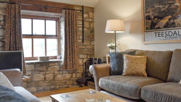 A cozy living room with a stone wall, a large window with curtains, a sofa with patterned cushions, a table with a glass bowl, and a lamp. - The Cottage at Wilson House (Barningham, near Richmond)