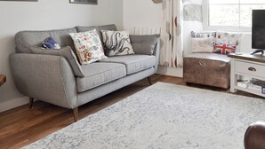 A living room with a grey sofa, patterned cushions, a white rug, a small ottoman, a white side table with a TV, and a window with curtains. - 3 bedroom accommodation in The Lizard Village, near Helston (The Lizard Village, near Helston)