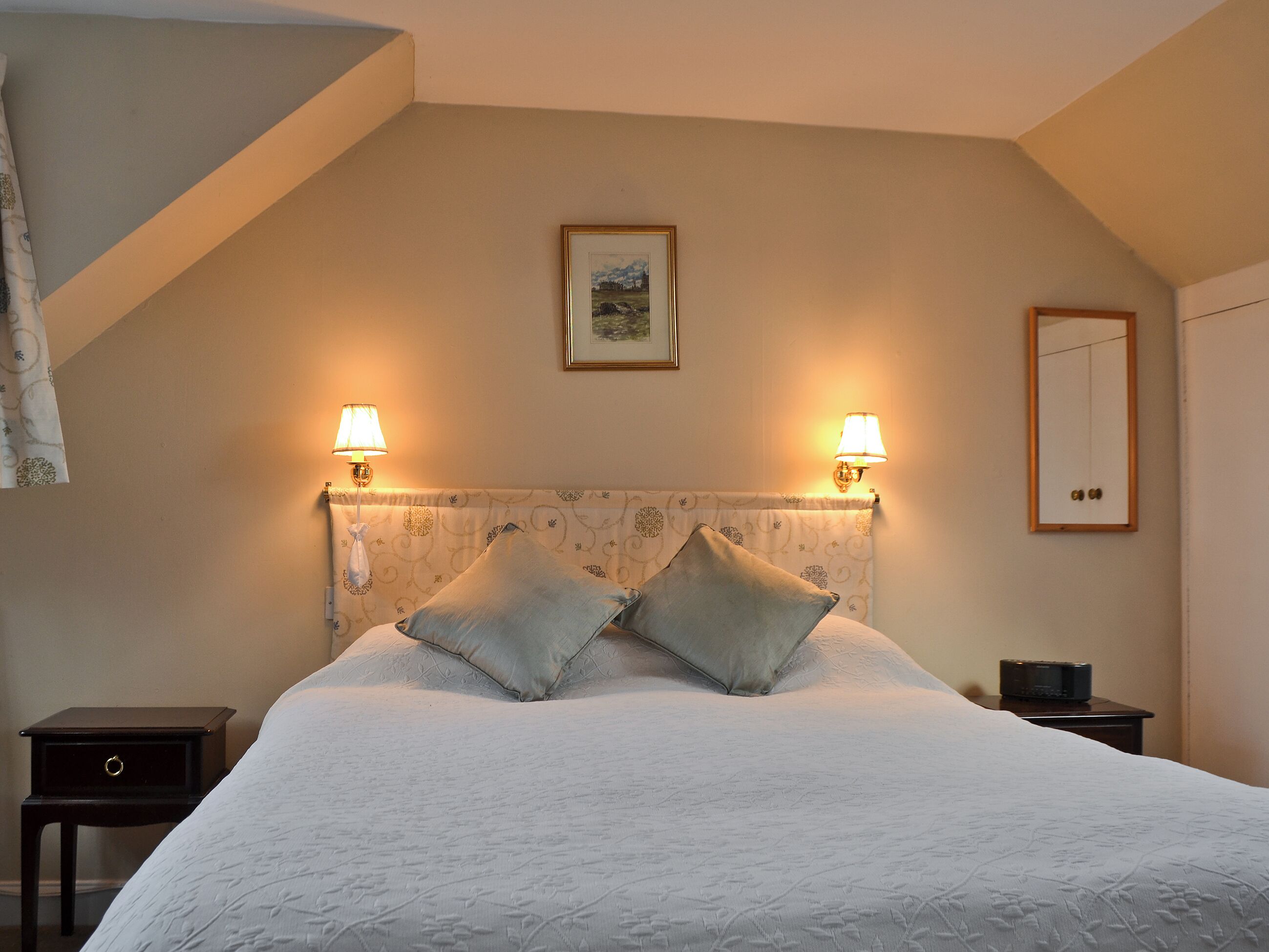 A bedroom with a large bed, two bedside lamps, a framed picture on the wall, and a mirror on the wall.
