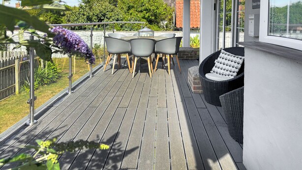Une terrasse en bois avec une table et des chaises, entourée d’un mur blanc et d’une porte vitrée.