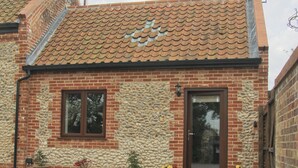 Een gebouw van baksteen met een pannendak, een stenen muur en een deur. Er staan potplanten ervoor.