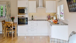 A modern kitchen with white cabinetry, a built-in oven, microwave, and a stainless steel range hood. There is a dining area with a wooden table and chairs, and a window with curtains. - 2 bedroom accommodation in Bodmin (Bodmin)