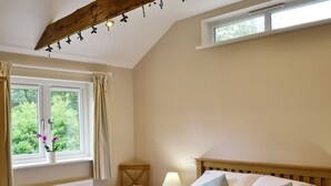A bedroom with a bed, a window with curtains, a chair, a nightstand with a lamp, and a radiator. - 2 bedroom accommodation in Bodmin (Bodmin)