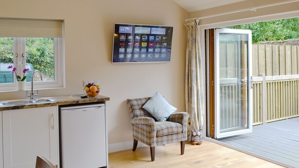 A compact kitchen with a built-in dishwasher, a dining area with a chair, and a large screen TV mounted on the wall. - 2 bedroom accommodation in Bodmin (Bodmin)