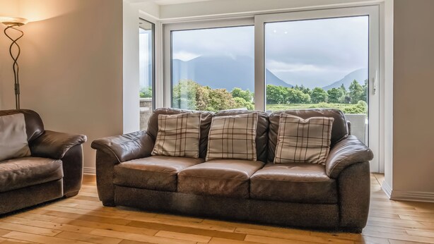 Una sala de estar con un sofá de cuero marrón, un sillón marrón y una lámpara de pie. La habitación tiene ventanas grandes con vistas a un paisaje montañoso.