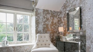 Een badkamer met een witte gestoffeerde stoel, een wastafel met spiegel en bloemenbehang.