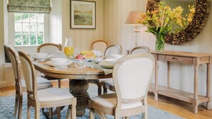 Een eetruimte met een ronde tafel, stoelen, een vaas met bloemen, een lamp en een decoratieve spiegel.