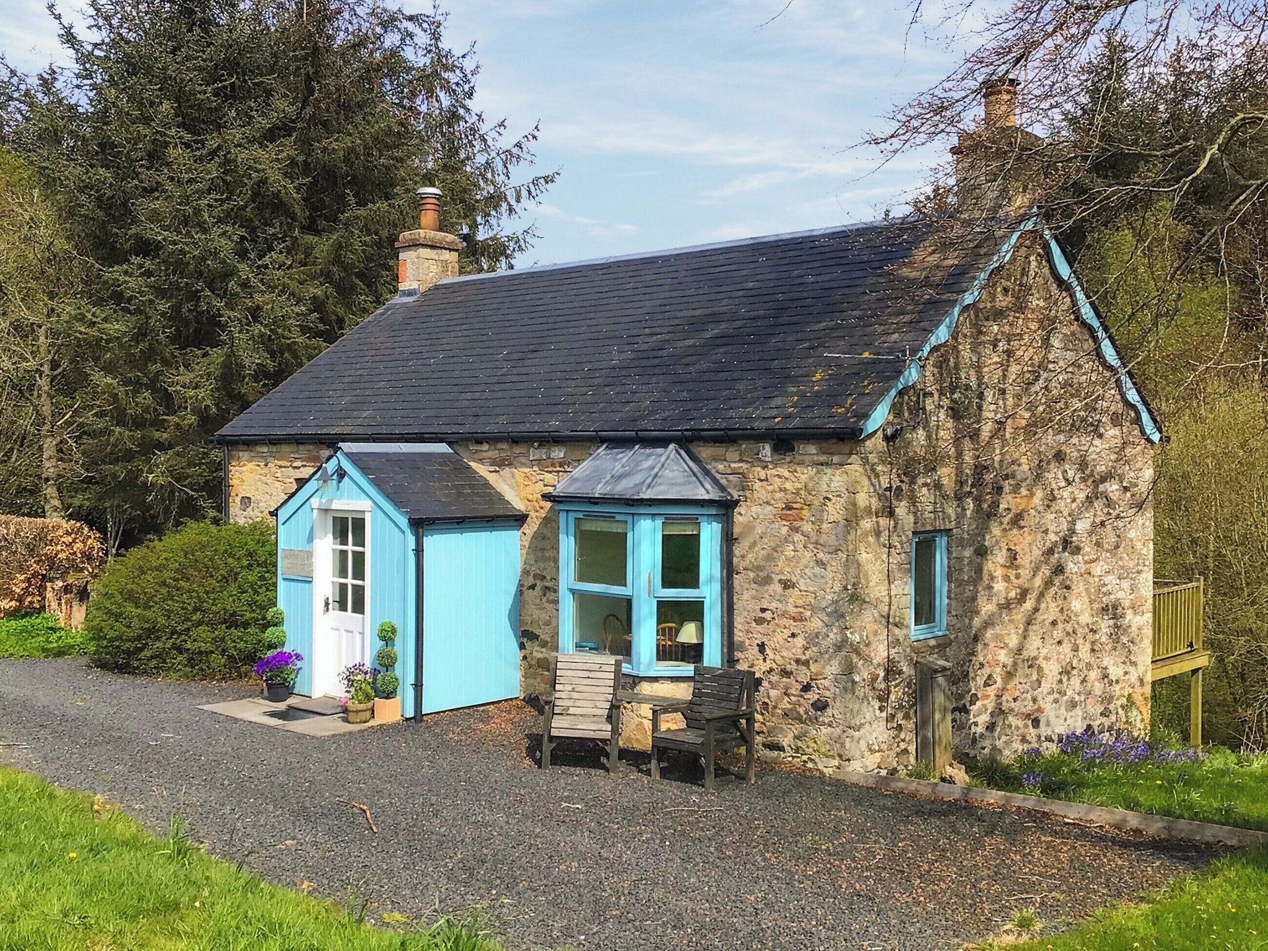 Une petite maison en pierre avec une porte et des fenêtres bleues, une cheminée et une allée gravillonnée.