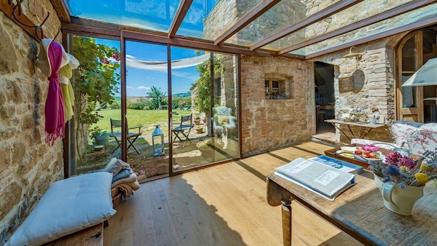 A cozy room with a stone wall, a wooden table with a newspaper, and a view of a garden through large glass doors. - VILLA MERAVIGLIOSA - Six Bedroom Villa, Sleeps 12 (Pienza)