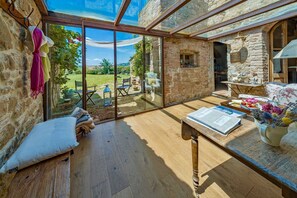 A cozy room with a stone wall, a wooden table with a newspaper, and a view of a garden through large glass doors. - VILLA MERAVIGLIOSA - Six Bedroom Villa, Sleeps 12 (Pienza)
