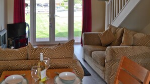 Una sala de estar con sofás estampados, una mesa de comedor con platos y copas de vino, un televisor y vistas a un jardín a través de puertas de cristal.