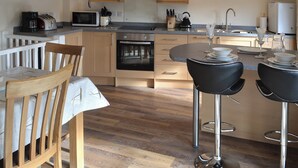 A modern kitchen with wooden cabinets, a built-in oven, a microwave, and a dining area with a table and chairs. - 2 bedroom accommodation in Port Isaac (Port Isaac)