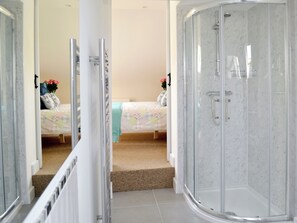 A bathroom with a glass shower enclosure, a bed with floral bedding, and a small sitting area with a chair and a plant. - 2 bedroom accommodation in Hartland, near Bideford (Hartland, near Bideford)