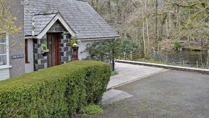 A stone building with a sloped roof, a wooden door, and a small garden area with a hedge and a hanging flower pot.
