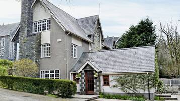 A two-story stone building with a stone chimney, a small attached structure with a tiled roof, and a well-maintained hedge.