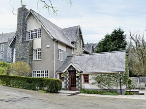 A two-story stone building with a stone chimney, a small attached structure with a tiled roof, and a well-maintained hedge.