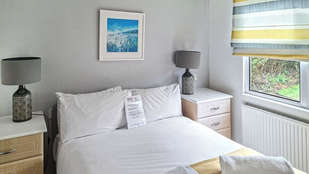 A neatly made bed with white linens, a bedside table with a lamp, a framed picture on the wall, and a window with a view of greenery. - 4 bedroom accommodation in East Runton, near Cromer (East Runton, near Cromer)