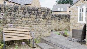 Un muro de piedra, un banco de madera, una silla de mimbre y un pequeño jardín con grava y plantas.