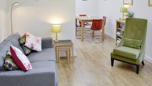 Una sala de estar con un sofá gris, una mesita con una lámpara, una mesa de comedor con sillas y un sillón verde.