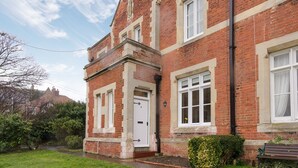 Une maison à deux étages, aux murs de briques rouges, avec une porte et des fenêtres blanches, un petit jardin à l’avant et un banc.