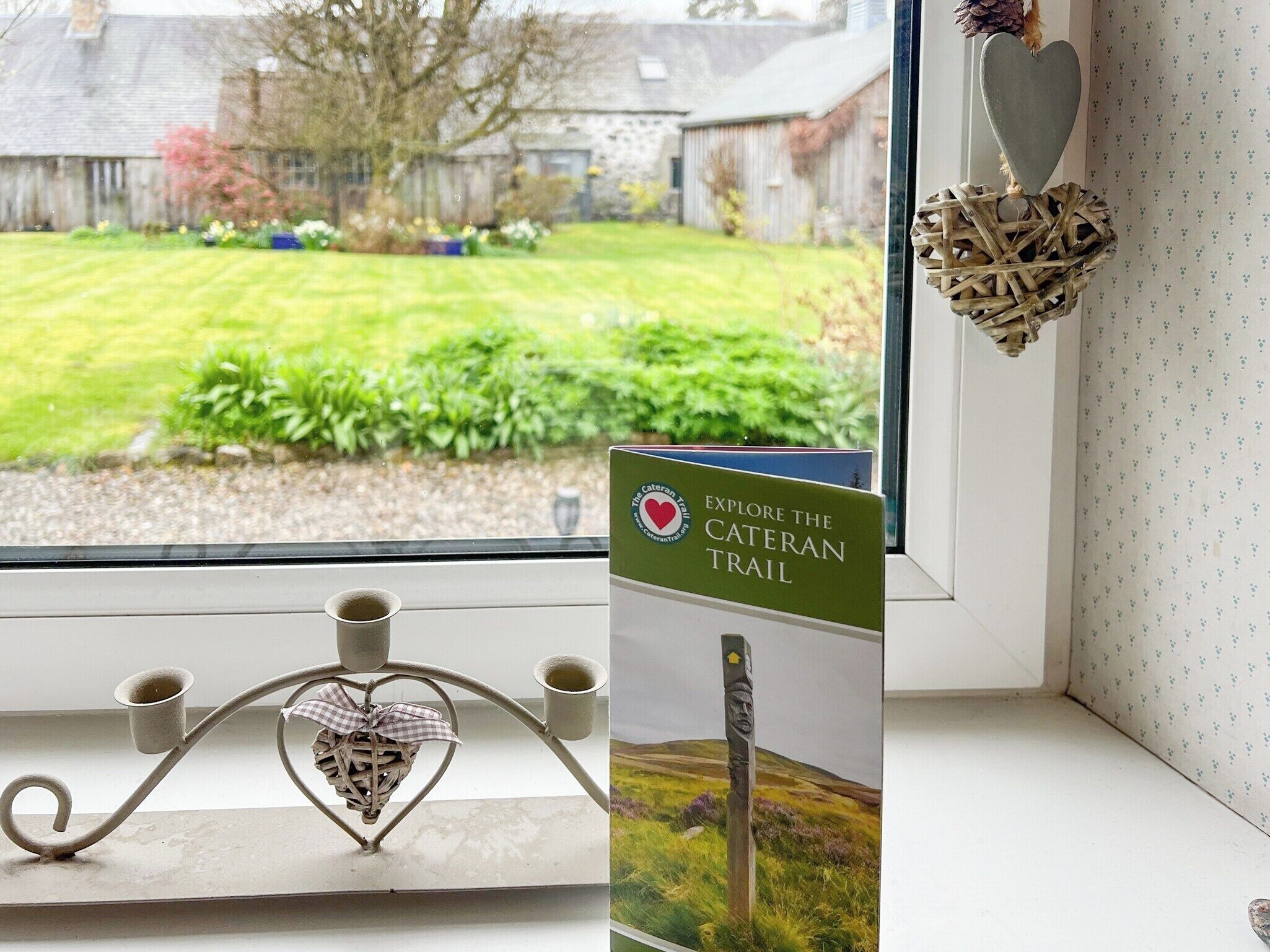 A window with a view of a green garden, a sign for the CATERAN TRAIL, and a decorative heart-shaped candle holder.