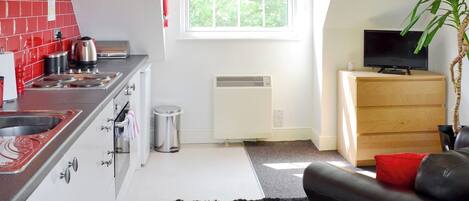 A compact kitchen with a stove, sink, and a small trash bin. There's a window with a red valance, a radiator, and a black sofa with a red pillow. A television is on a wooden cabinet.