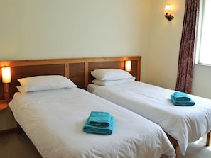 Une chambre d’hôtel avec deux lits simples, chacun doté d’une tête de lit en bois et de linge de lit blanc. Une lampe fixée au mur se trouve sur le côté gauche du lit, et une fenêtre avec des rideaux se trouve sur le côté droit.