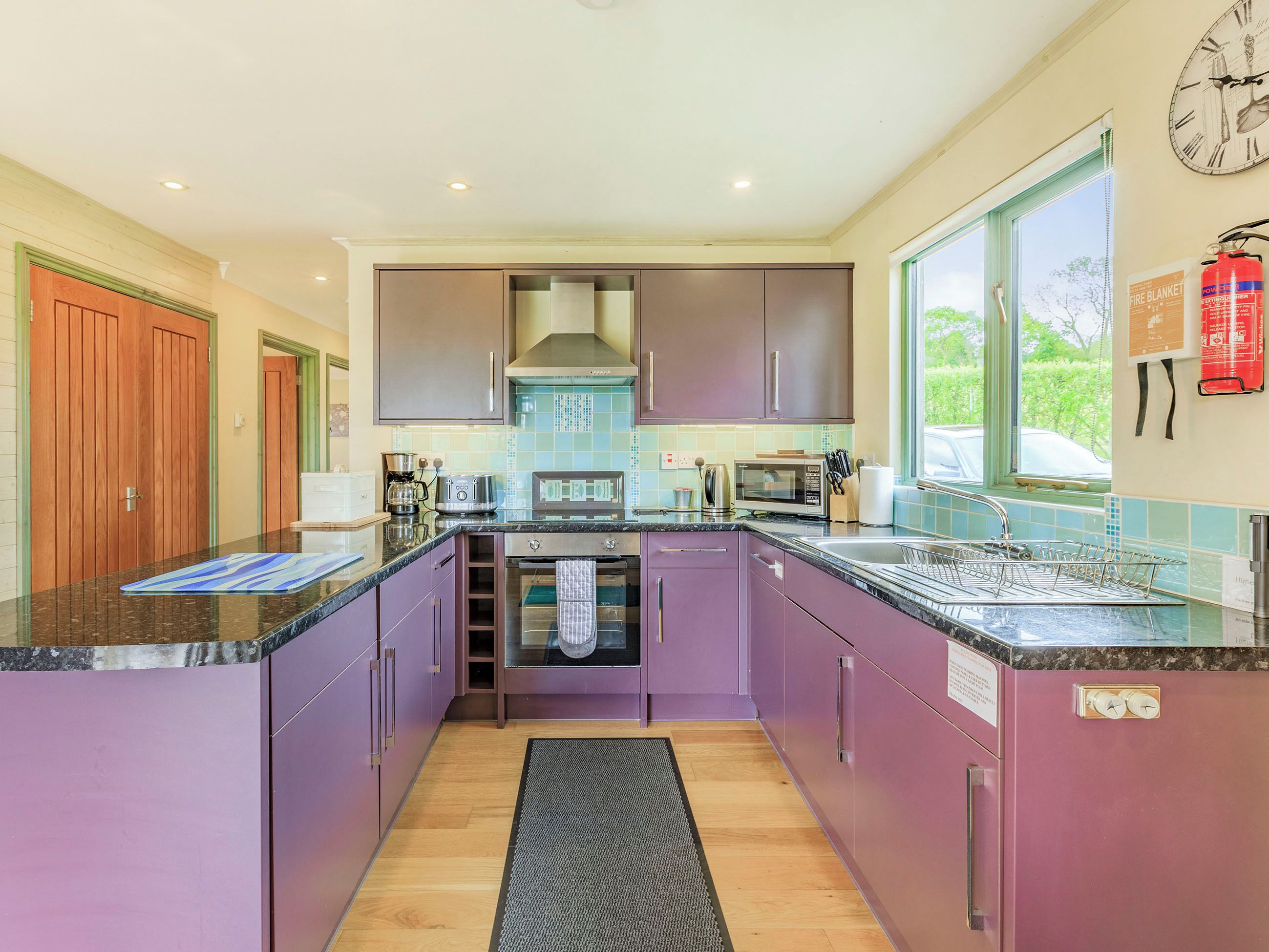 A modern kitchen with purple cabinets, stainless steel appliances, and a large window providing a view of the outdoors.