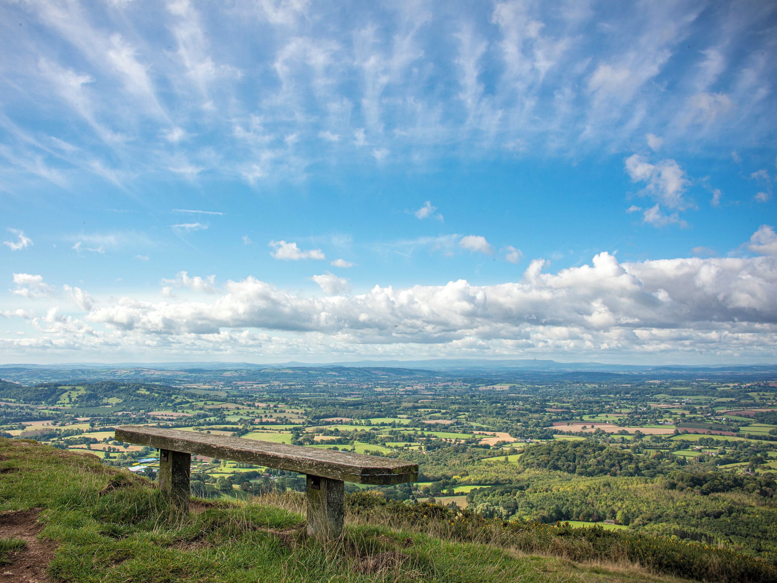 A stone bench overlooking a vast landscape with rolling hills and fields under a partly cloudy sky.