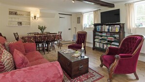 A living room with a wooden floor, a dining table with chairs, a red velvet armchair, a bookshelf, and a television on the wall. - 3 bedroom accommodation in Denford, near Thrapston (Denford, near Thrapston)