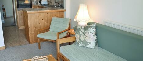 Een knus woongedeelte met een kitchenette, een bank, een fauteuil, een lamp en een tijdschrift op een salontafel.