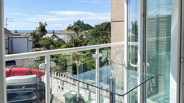 Un balcone con tavolo e sedie in vetro, affacciato su una zona costiera con edifici e un castello.